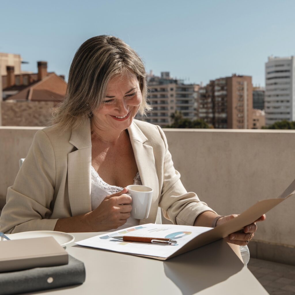 Una mujer vestida de negocios está sentada en una mesa al aire libre, sosteniendo una taza de café y revisando documentos con gráficos