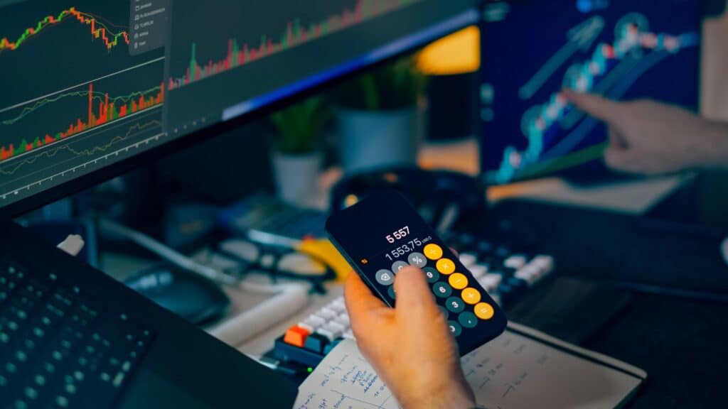 Persona usando calculadora en el celular mientras analiza gráficos financieros en múltiples pantallas.