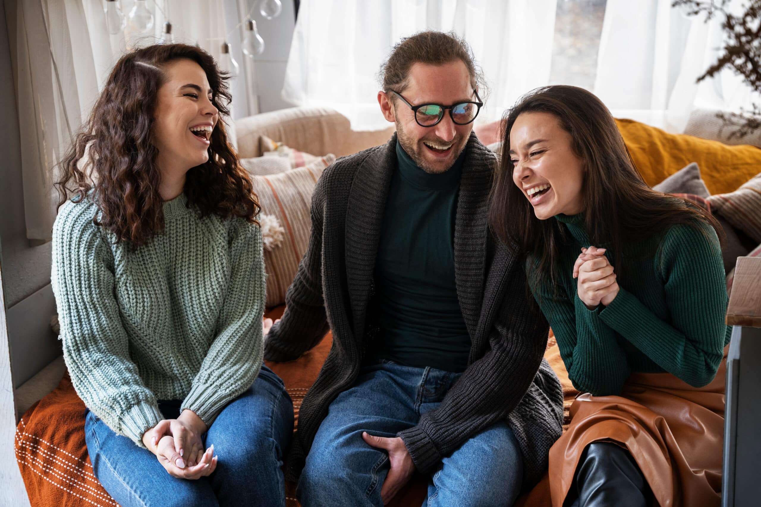 Tres personas están sentadas en un sofá, sonriendo y riendo juntas en una habitación acogedora y bien iluminada, con almohadas y mantas.