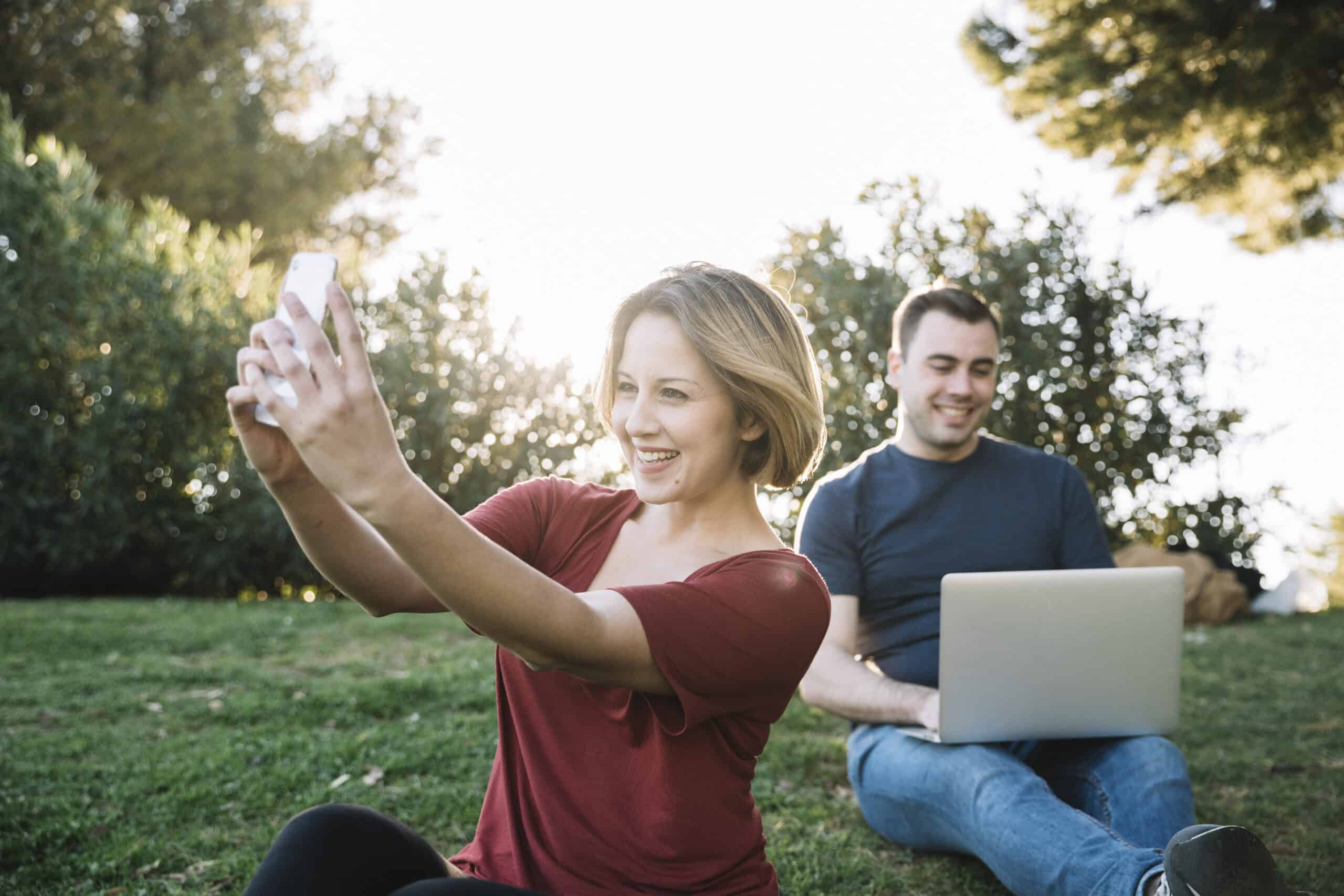 Mujer tomándose una selfie con su celular en un parque mientras otra persona usa un computador portátil al aire libre.