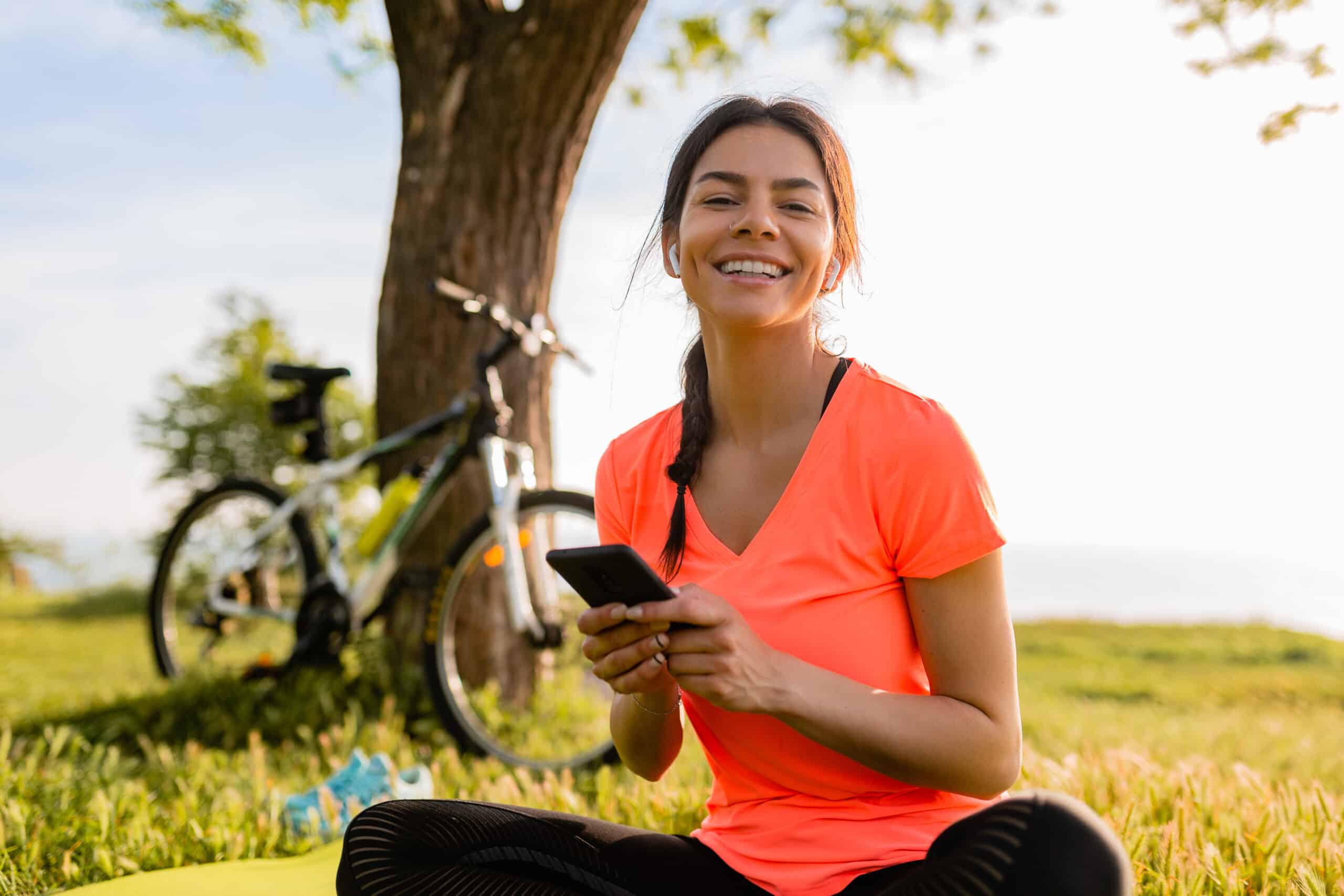 Mujer sonriente sentada en un parque usando su celular después de hacer ejercicio, con una bicicleta al fondo.