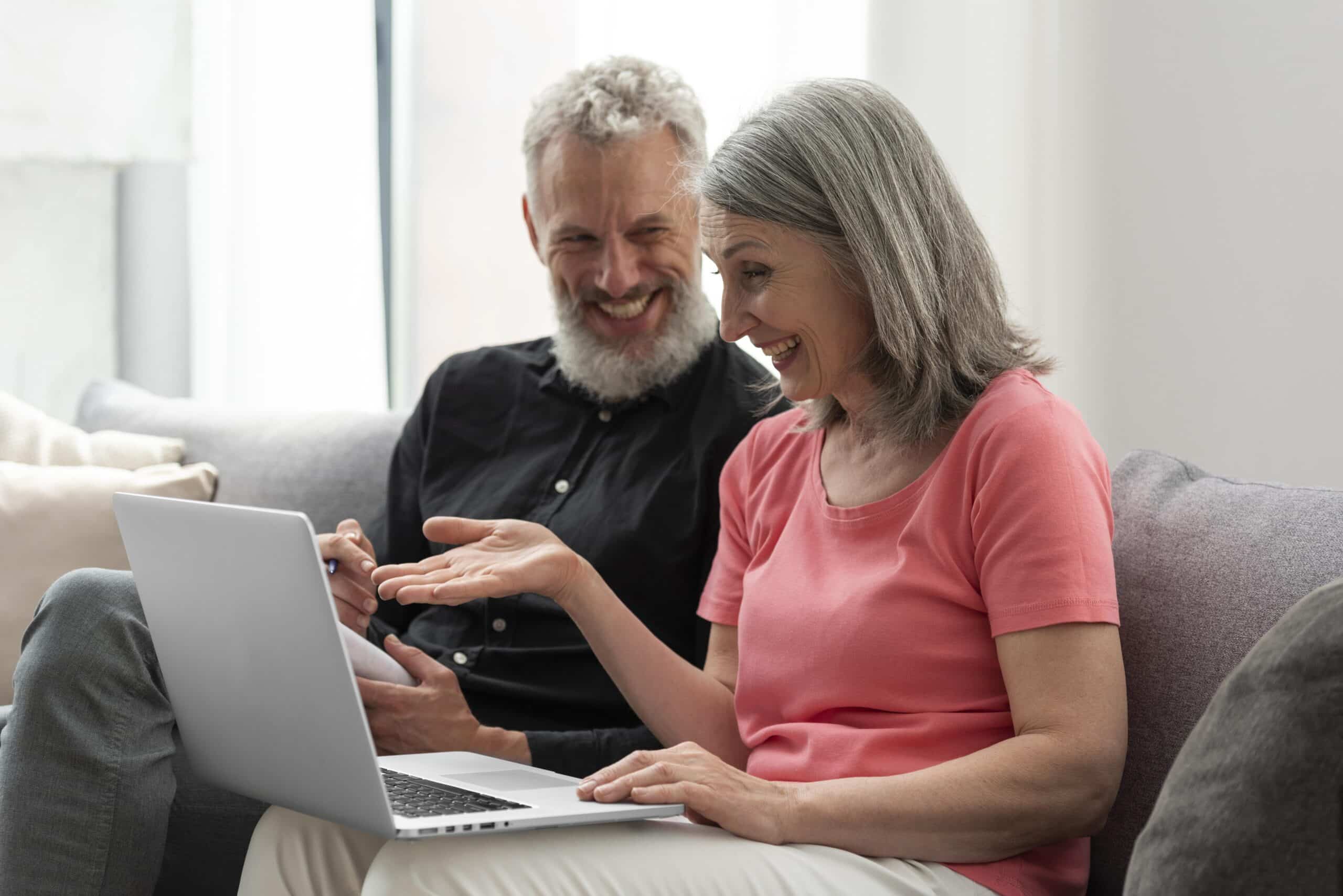 Pareja mayor sentada en un sofá, usando una computadora portátil y sonriendo juntos